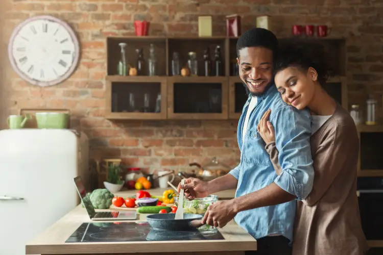 black husband preparing food in kitchen while black wife hugging from back side